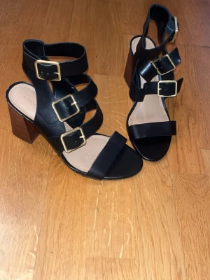 Svarta sandaler med klack och spännen - Stilrena svarta sandaler med breda remmar och fyra stora guldfärgade spännen. Skorna har en öppen tå och en stabil, brun träliknande blockklack. Tillverkade i syntetmaterial med mjuk innersula för extra komfort.