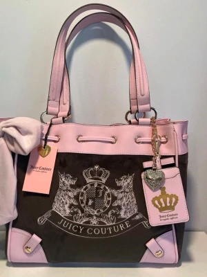 Juicy Couture daydreamer scottie brun och rosa tote - Stilren handväska från Juicy Couture i brun mocka med ljusrosa detaljer i skinnimitation. Väskan har dubbla axelband, dekorativ rosett, emblem med lejon och krona samt ett hängande hjärta och kortfack med guldkrona. Perfekt för dig som gillar en trendig och lyxig look.