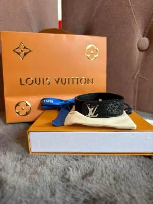 Louis Vuitton svart läderarmband - Snyggt svart armband från Louis Vuitton i äkta läder med silverfärgat LV-logo framtill. Armbandet har klassiskt monogrammönster och stängs med spänne. Perfekt accessoar för att lyfta din outfit med en lyxig touch.