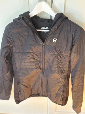 Svart dunjacka från 8848 Altitude - Snygg svart dunjacka från 8848 Altitude med huva och dragkedja framtill. Jackan har diskret quiltad design, två fickor med dragkedja och en vit 8-logga på bröstet. Perfekt för kalla dagar och enkel att matcha med olika outfits.
