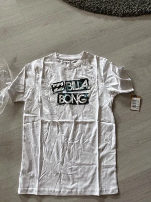 Vit Billabong t-shirt med tryck - Vit t-shirt från Billabong med ett stort grafiskt tryck i svart, blått och vitt på bröstet. Storlek xs , aldrig använd. Nypris 210kr