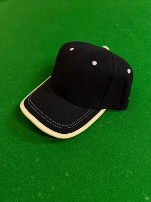 Svart keps med vit kant - Snygg svart keps med vit kant runt skärmen och vita detaljer på ovansidan. Klassisk böjd skärm och justerbar passform baktill. Perfekt accessoar för en sportig och avslappnad stil.