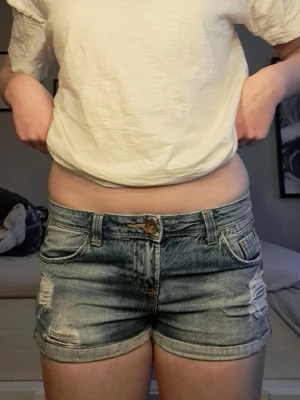 Blå slitna jeansshorts från Denim Factory - Snygga blå jeansshorts från Denim Factory med slitna detaljer och klassisk femficksdesign. Lågmidjade med uppvikta benslut och knappgylf. Perfekta för sommaren och lätta att matcha med allt.