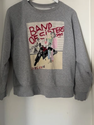 Grå sweatshirt Zadig & Voltaire - Grå sweatshirt från Zadig & Voltaire x Band of Sisters med tryck fram och texten 'BAND OF SISTERS SAYS' samt illustration av två tjejer och motorcykel. På ryggen står det 'SISTERHOOD IS POWERFUL'. Mjuk bomullskänsla och avslappnad passform.