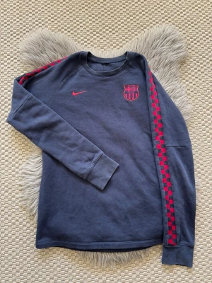 Blå Nike FC Barcelona hoodie - Säljer en blå hoodie från Nike med FC Barcelona-logga och röd Nike-symbol på bröstet. Ärmarna har ett rött rutmönster längs sidorna. Tröjan har ribbade muddar och är tillverkad i mjukt material, perfekt för dig som gillar sportiga och snygga kläder.