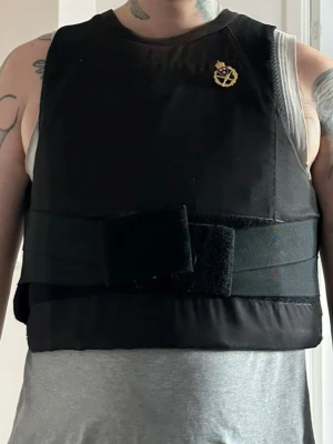 SAFEGUARDARMOUR/ballistisk/Skottsäker/väst, svenskt skyttemärke/försvarsmakten - Denna väst är designad för att bäras över andra plagg och har en justerbar passform med kardborreband i midjan. Den är utrustad med ett svenskt skyttemärke som indikerar att bäraren har uppnått en viss nivå inom gevärsskytte, ofta inom militär eller organiserad skytteverksamhet. Märket har de tre kronorna som är rikssymbolen för Sverige. Den skyddar mot kniv och alla pistoler. Den är XL men killen på bilden har M så alla som har M/L/XL kan ha den. Obs. Patronerna ingår ej.