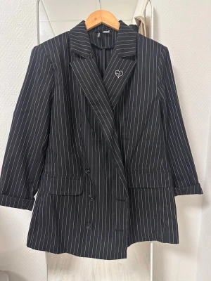 Svart randig kavaj med nål H&M - Snygg svart kavaj från H&M med vita tunna ränder och klassisk krage. Kavajen har dubbelknäppning, två fickor framtill och en dekorativ nål i form av ett hjärta på kragen. Perfekt för en trendig och stilren look.