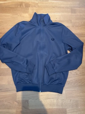  Mörkgrlå zip hoodie från Fred Perry - Snygg mörkgrå hoodie från Fred Perry med dragkedja hela vägen och hög krage. Klassisk design med ribbade muddar och diskret logga på bröstet. Perfekt för en stilren och sportig look. Storlek M men skulle vilja påstå att det egentligen är S. Köpt från NK Göteborg för ca 1600. Har tyvärr tappat bort kvittot.