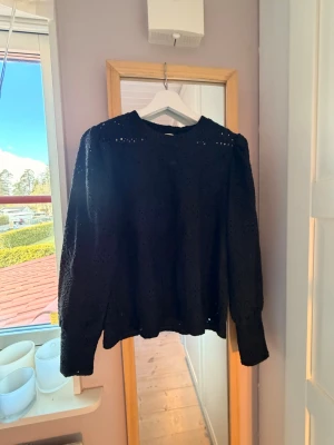 Svart spetsblus med lång ärm - Snygg svart blus med lång ärm och broderad spetsdetalj. Blusen har rund halsringning och ett genombrutet mönster som ger en cool och trendig look. Perfekt att styla med jeans eller kjol för en uppdaterad outfit.