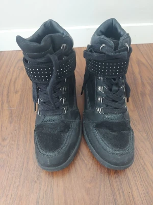 Svarta high top sneakers med nitar - Snygga svarta high top sneakers med snörning och bred kardborrem över vristen dekorerad med små silverfärgade nitar. Skorna har en mix av mocka och textilmaterial samt robust sula. Perfekta för dig som vill ha en edgy och trendig look.