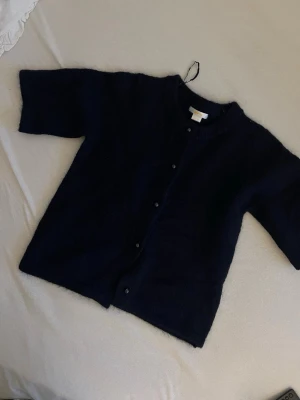 Mörkblå kofta från Gina Tricot - Mysig mörkblå kofta från Gina Tricot med rund halsringning och knappar framtill. Koftan har en fluffig, stickad känsla och halvlånga ärmar. Perfekt att slänga på sig över en t-shirt för en chill look.