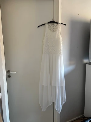 Vit långklänning med spetsdetaljer - Snygg vit långklänning med broderad spets upptill och flowy, asymmetrisk kjol. Ärmlös modell med rund halsringning och lätt transparent nederdel. Perfekt för sommarens alla tillfällen.