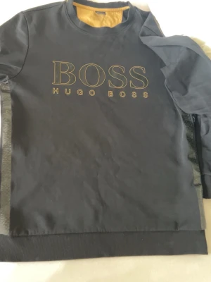 Svart sweatshirt från Hugo Boss - Svart sweatshirt från Hugo Boss med stor guldfärgad logga på bröstet. Tröjan har rund halsringning och ribbade muddar vid ärmslut och nederkant. Perfekt för en stilren och avslappnad look.
