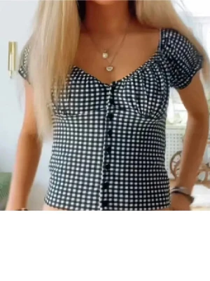 Rutig topp från Stradivarius - Supersöt svartvit rutig topp från Stradivarius med knappar framtill och korta puffärmar. Figurnära passform och v-ringad halsringning. Perfekt för dig som gillar trendiga och feminina plagg.