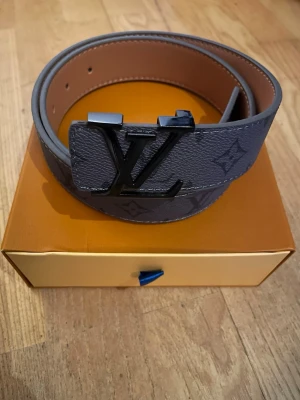  mörkgrå bälte - Snyggt mörkgrått bälte med klassiskt spänne i svart. Bältet har det ikoniska monogrammönstret. Perfekt accessoar för att lyfta din outfit.