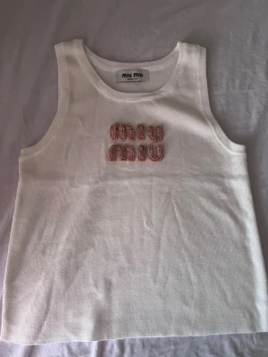 Vit linnetopp från Miu Miu med pärlor - Superfin vit linnetopp från Miu Miu med broderad logga framtill i rosa pärlor och stenar. Toppen är ärmlös och har en klassisk rund halsringning. Perfekt för dig som vill ha något stilrent men ändå extra.