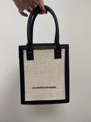 Beige/svart handväska CHAU NGTAI&OBI - Stilren handväska i beige canvas med svarta detaljer och handtag. Väskan har en fyrkantig form och svart ram i konstläder samt avtagbar axelrem. Märket CHAU NGTAI&OBI står tryckt på framsidan. Perfekt för dig som gillar minimalistisk och trendig design.