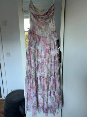 Blommig långklänning från Anaya - Romantisk långklänning från Anaya med volanger. Klänningen har ett blommigt mönster i pastelliga toner av rosa, lila, orange och grönt på vit botten. Tunt, svepande tyg i polyester ger en luftig känsla. Står ingen storlek men XS/S.