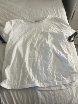 Vit t-shirt med svarta detaljer - Vit t-shirt med korta ärmar och svarta spetsdetaljer vid ärmsluten. Enkel och stilren design som passar till det mesta. Tillverkad i mjuk bomull för en skön känsla hela dagen. Har en liten maskara fläck fram.