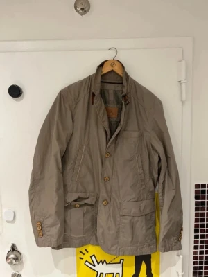 Beige rock från Massimo Dutti - Stilren beige rock från Massimo Dutti med klassisk krage och snygga knappar framtill. Rocken har flera praktiska fickor och detaljer i brunt läder vid kragen. Perfekt för dig som gillar en tidlös och avslappnad look.