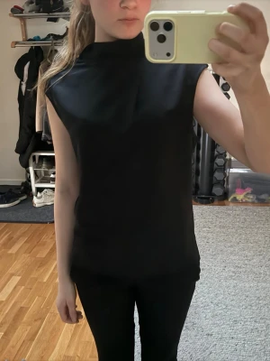 Svart ärmlös topp med hög hals - Stilren svart topp med hög hals och ärmlös design. Toppen har en rak passform och är perfekt att matcha med både jeans och kjol. Materialet känns mjukt och följsamt mot huden.
