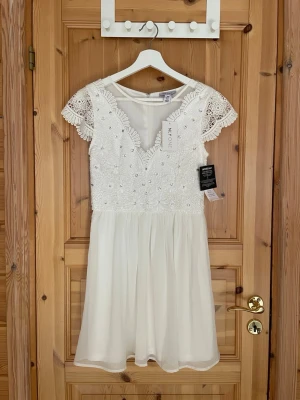 Vit spetsklänning från Nelly - Superfin vit kortklänning från Nelly med spetsdetaljer, pärlor och glittriga stenar på överdelen. Klänningen har v-ringning, korta ärmar i spets och en flowy kjol i tunt material. Perfekt för student eller sommarens alla tillfällen!☀️🎉
