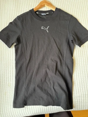 Svart Puma t-shirt med logga - Snygg svart t-shirt från Puma med klassisk logga broderad på bröstet. Tillverkad i ett strukturerat, sportigt material som andas. T-shirten har rund halsringning och korta ärmar, perfekt för en aktiv stil.