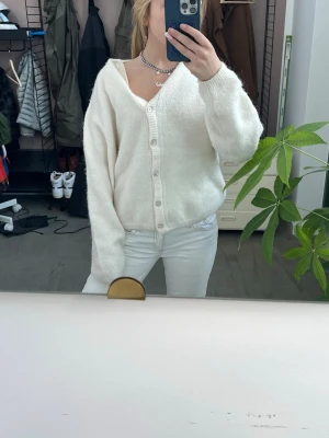 Vit fluffig kofta med knappar - Mysig vit kofta med fluffig känsla. Koftan har långa ärmar och stängs med knappar framtill. Perfekt att slänga på sig över en topp för en avslappnad look.