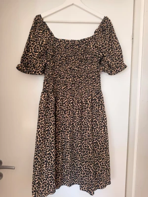 Leopardmönstrad klänning med puffärm - Trendig leopardmönstrad klänning med smockad överdel och puffiga korta ärmar. Klänningen har en fyrkantig ringning och är tillverkad i ett mjukt, lätt tyg som ger en snygg siluett. Perfekt för dig som vill sticka ut med ett djurmönster.