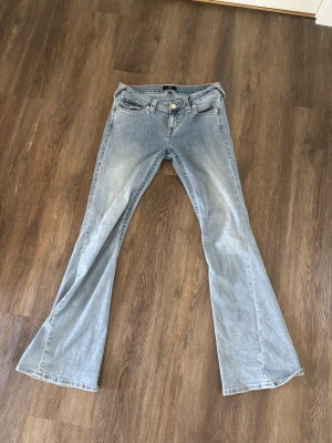 Ljusblå bootcut jeans från true religion  - Snygga ljusblå bootcut jeans från true religion med klassisk femficksdesign och utsvängda ben. Jeansen har en vintage tvätt och detaljerade bakfickor med knappar. Perfekta för en avslappnad och trendig look. Stolek 28, och modell joey, knappt använda🥰