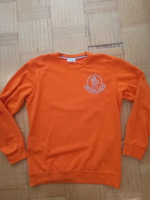 Orange sweatshirt från Moncler - Snygg orange sweatshirt från Moncler med rund halsringning och stort vitt logotryck på bröstet. Tröjan har långa ärmar och ribbade muddar vid ärmslut och nederkant. Perfekt för dig som vill sticka ut med färg. Storleken säger XXL men den passar även XL