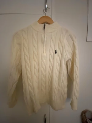 Vit kabelstickad tröja Polo Ralph Lauren - Kabelstickad vit tröja från Polo Ralph Lauren med halv dragkedja och klassisk broderad logga på bröstet. Tröjan har ribbade muddar och hög krage, perfekt för lager på lager. Materialet känns mjukt och kvalitativt.