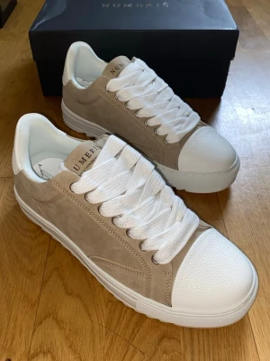 Beige och vita sneakers från Numeris - Stilrena sneakers från Numeris i beige mocka med vita detaljer och breda vita skosnören. Skorna har en klassisk låg profil, vit gummisula och rund tå. Perfekta för dig som gillar en clean och modern look.
