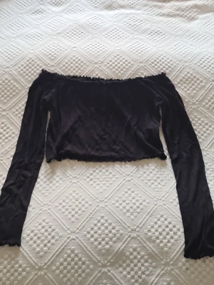 Svart offshoulder topp från Bik Bok - Svart offshoulder topp från Bik Bok med långa ärmar och croppad passform. Toppen har en lätt ribbad struktur och små volangdetaljer vid kanterna.
