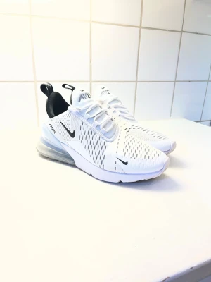 Nike Air Max 270 vita sneakers - Nike Air Max 270 sneakers i vitt med svarta detaljer och stor synlig Air-enhet i hälen. Ovandelen är i mesh för bra ventilation och har en modern, sportig look. Klassisk Nike Swoosh på sidan och dragflik bak för enkel påtagning.