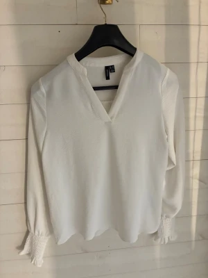Vit blus med v-ringning från veromoda - Stilren vit blus från veromoda med v-ringad hals och långa ärmar. Ärmsluten har bred smock och volangkant som ger en trendig touch. Blusen är tillverkad i ett lätt, mjukt tyg som faller snyggt och passar perfekt till både jeans och kjol.