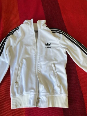 Vit Adidas track jacket - Säljer en klassisk vit Adidas track jacket med svarta ränder längs ärmarna och Adidas logga på bröstet. Jackan har dragkedja framtill, två fickor med dragkedja och ribbade muddar. Perfekt för en sportig och avslappnad stil.
