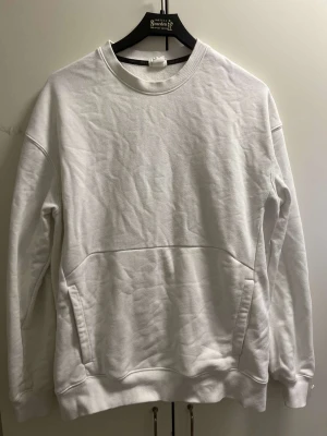Vit sweatshirt utan huva - En stilren vit sweatshirt från HM med rund halsringning och ribbade muddar vid ärmslut och nederkant. Tröjan har en stor magficka framtill och är tillverkad i mjukt bomullsmaterial. Perfekt för en avslappnad och trendig look.