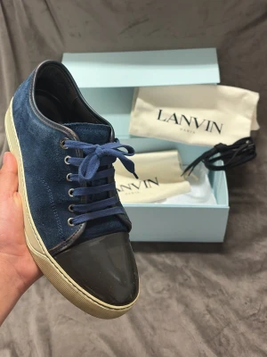 Blåa Lanvin sneakers med mocka och lack - Snygga sneakers från Lanvin i blå mocka med svart lackad tå och vita sulor. Skorna har blå skosnören och klassisk låg profil. Perfekta för dig som gillar stilrena och exklusiva sneakers med unika materialval.