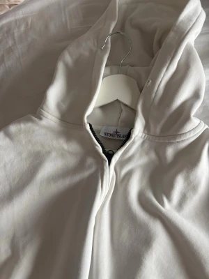 Vit hoodie från Stone Island - Snygg vit hoodie från Stone Island med dragkedja och huva. Tillverkad i mjukt bomullsmaterial som känns skönt mot huden. Perfekt för en clean och stilren look. Klassisk passform och diskret logga i nacken.