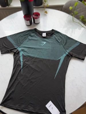Svart träningströja från Gymshark V5 - Svart kortärmad träningströja från Gymshark med turkosa grafiska detaljer över bröstet och axlarna. Tillverkad i ett lätt och snabbtorkande material som passar perfekt för intensiva träningspass. Gymshark-logga framtill.
