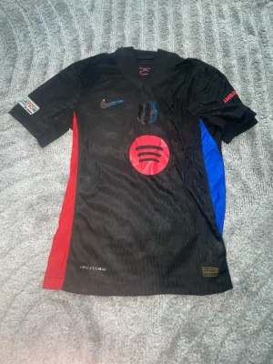 Svart FC Barcelona matchtröja Nike - Säljer en svart FC Barcelona matchtröja från Nike med klubbmärke på bröstet, röd och blå detalj på sidorna samt Spotify-logga framtill. Tröjan har korta ärmar och är tillverkad i lätt funktionsmaterial, perfekt för fotbollsträning.