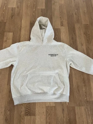 Grå Essentials Fear of God hoodie - Snygg ljusgrå hoodie från Essentials Fear of God med svart logga på bröstet. Klassisk passform med stor huva och magficka. Tillverkad i mjukt bomullsmaterial som känns riktigt skönt mot huden. Perfekt för dig som gillar streetwear och enkel stil.