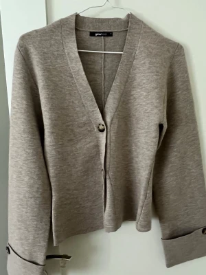 Beige kofta från Gina Tricot - Snygg beige kofta från Gina Tricot med v-ringning och två knappar framtill. Koftan har långa ärmar med dekorativa knappar vid ärmsluten och är gjord i ett mjukt, stickat material. Perfekt att slänga över en t-shirt eller topp.