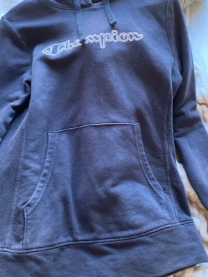 Svart Champion hoodie med logga - Svart hoodie från Champion med vit broderad logga på bröstet. Klassisk känguruficka framtill och justerbar huva med dragsko. Mjuk bomullsblandning som är skön att ha på sig. Perfekt för en avslappnad och sportig stil.