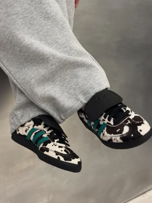 Adidas sneakers med koskinnsmönster - Unika Adidas sneakers med svartvitt koskinnsmönster och gröna ränder på sidorna. Skorna har svarta snören, bred svart plös med Adidas-logga och svart sula. Materialet ser ut att vara konstläder eller mocka med pälsstruktur. Perfekt för dig som vill sticka ut!