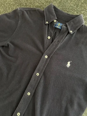 Mörkblå skjorta från Ralph Lauren - Klassisk mörkblå skjorta från Ralph Lauren med button-down krage och vita knappar. Skjortan har det ikoniska broderade logotypen på bröstet och är tillverkad i mjuk bomull. Perfekt för en stilren och avslappnad look.