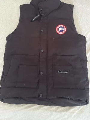 Svart dunväst från Canada Goose - Svart dunväst från Canada Goose med hög krage, två stora fickor framtill och klassisk logotyp på bröstet. Västen har knäppning med knappar och är tillverkad i slitstarkt material. Perfekt för kalla dagar och snygg att kombinera med hoodie eller t-shirt.