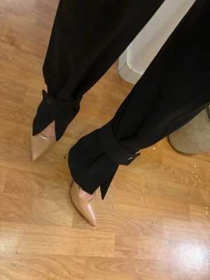Svarta kostymbyxor med knyt från H&M - Svarta kostymbyxor från H&M i storlek 36. Byxorna har vida ben och coola knytband med knappdetalj vid anklarna. Perfekta för en trendig och stilren look. Materialet känns mjukt och följsamt.
