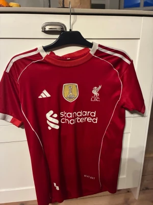 Liverpool matchtröja röd Adidas - Säljer en röd Liverpool matchtröja från Adidas med vita detaljer och klubbmärke på bröstet. Tröjan har FIFA-märke, sponsorlogga och korta ärmar. Tillverkad i funktionsmaterial som andas, perfekt för fotbollsträning eller match. Använd ett par gånger och har storleken L
